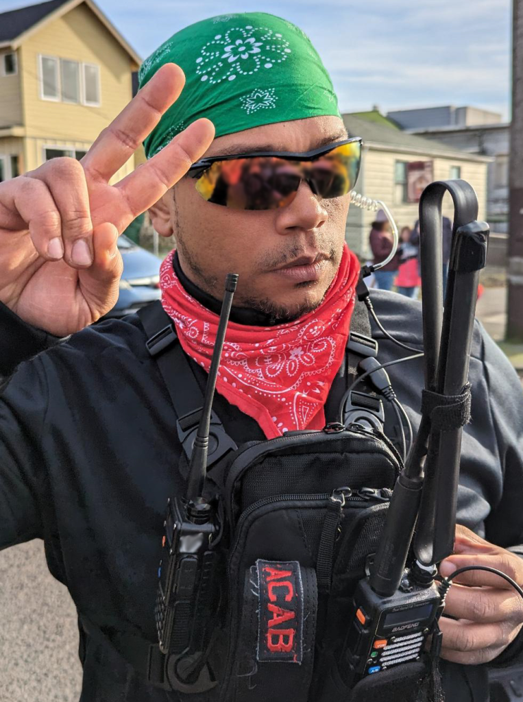 Light brown person of color throwing up a peace sign with their right hand, wearing highly reflective sunglasses with a yellow tint, two bandanas (green on head and red around neck), a black long sleeve shirt, and a black chest pouch with an "ACAB" patch attached sideways on a front pocket and two two-way-radios attached to the pouch. In the background is a neighborhood set under a cloudy blue sky.